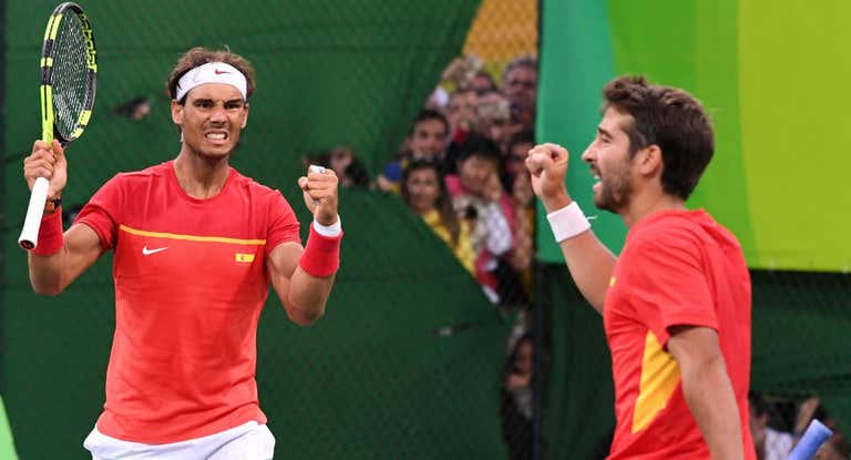Rafa Nadal y Marc López durante la semifinal del dobles olímpico en Río 2016. Al fondo, un grupo de aficionados siguiendo el partido desde fuera de la pista.